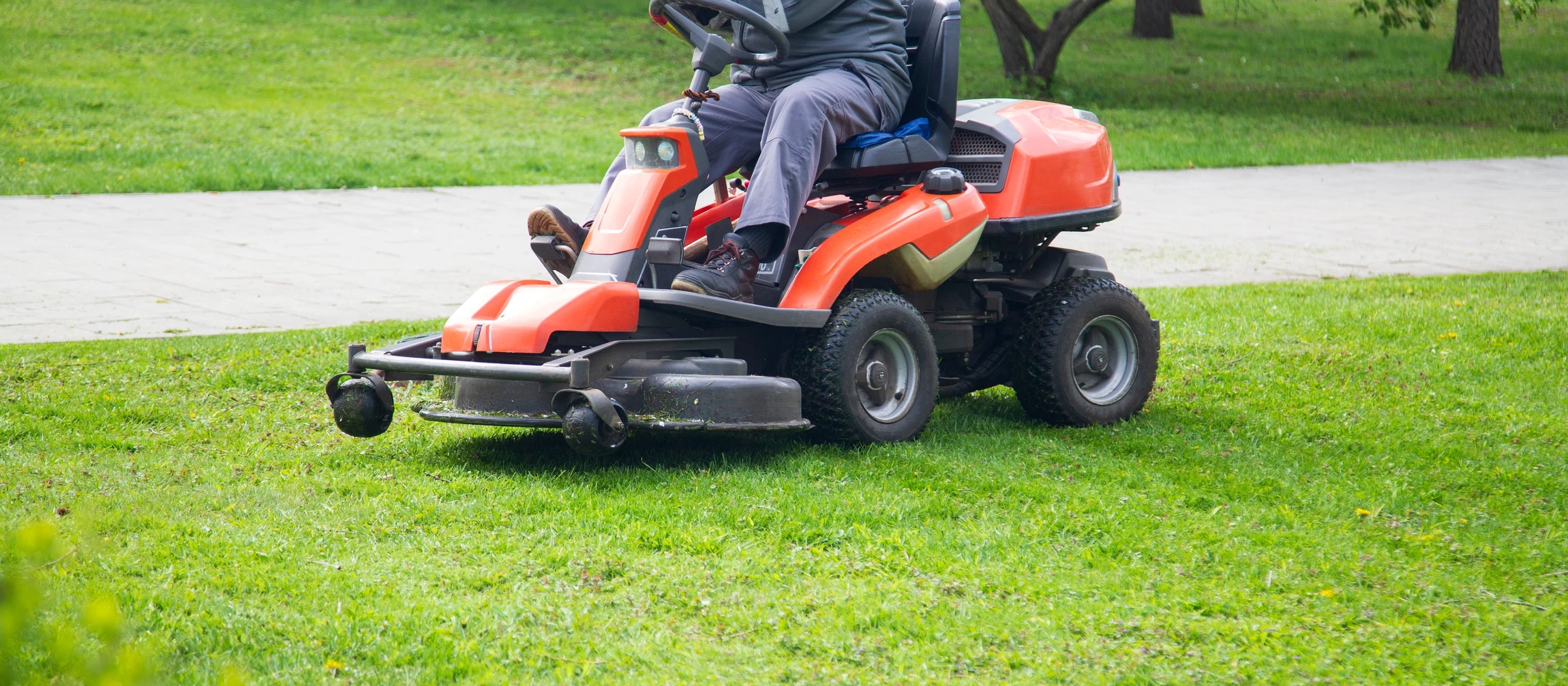 Scène extérieure d'entretien d'espaces verts avec opérateur sur tondeuse autoportée professionnelle orange effectuant la tonte d'une pelouse bien entretenue